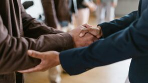 Two people shaking hands, both in long sleeve shirts—one navy blue and one brown—after discussing a business acquisition.