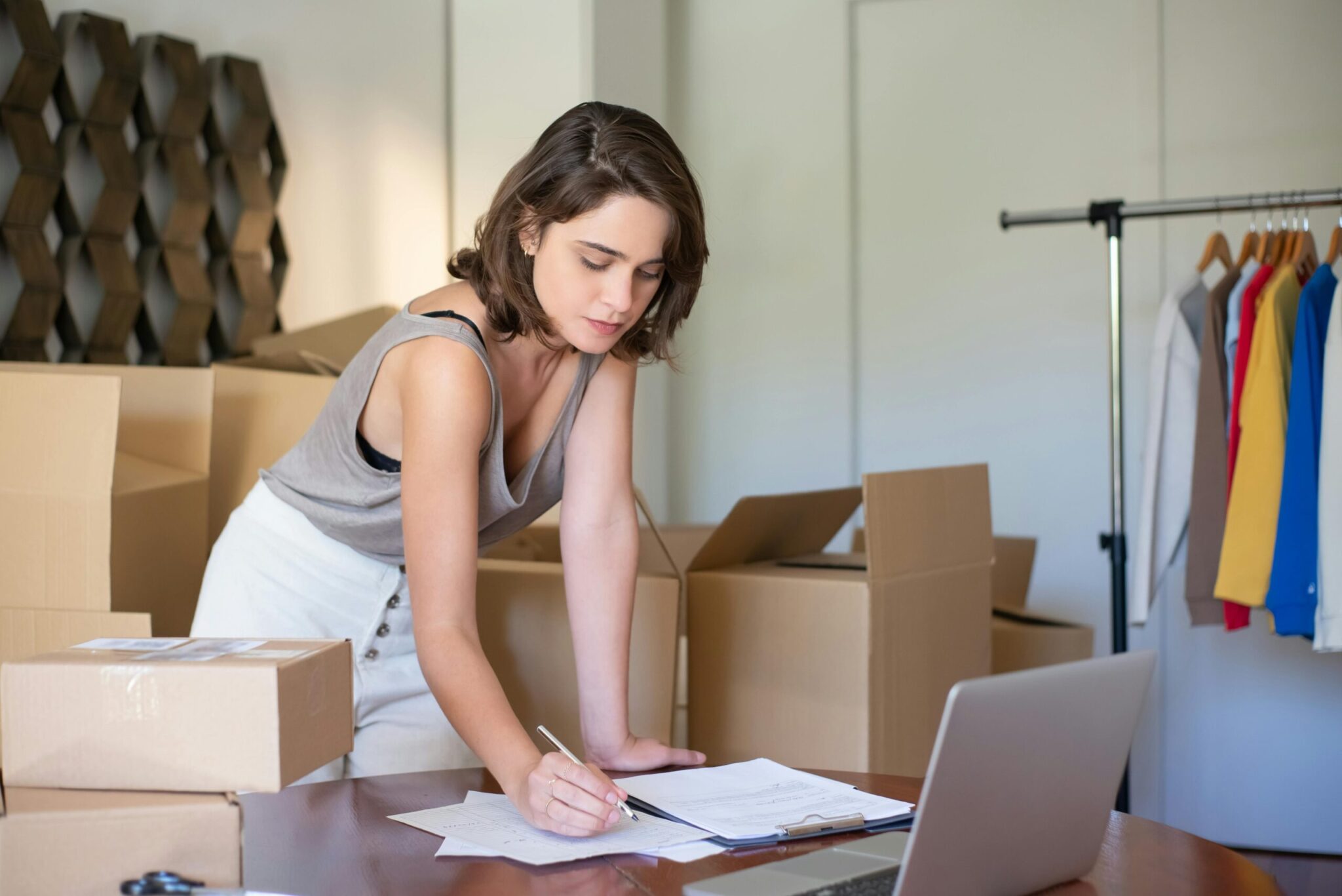 A woman stands among cardboard boxes, writing on a piece of paper at a desk with a laptop, symbolizing self-employed retirement planning.